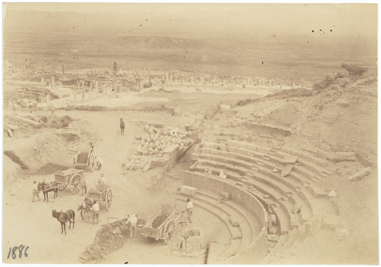 A sepia photograph from 1886 showing Timgad's theatre during excavation, with horse-drawn carts and stacked stone blocks in the foreground and the ruins of the city spreading toward the horizon.