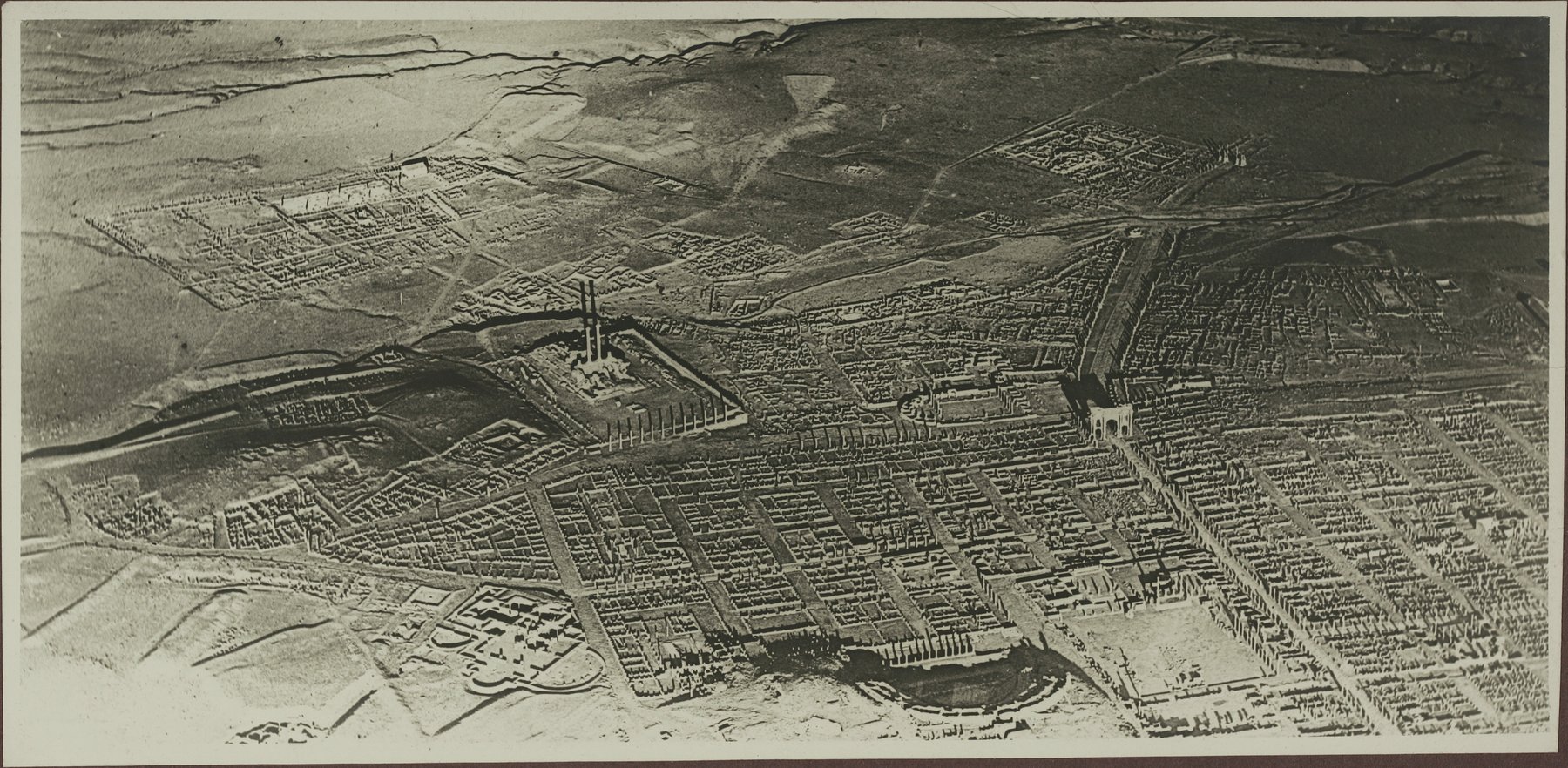 An aerial black and white photograph of Timgad taken in 1922 showing the rectilinear grid of insulae, the Capitol columns rising above the forum area, and the semicircular theatre visible at the bottom centre. The excavated city contrasts with the open ground surrounding it.