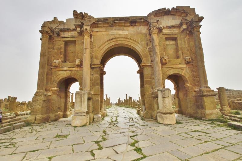 A colour photograph of the three-bay Arch of Trajan at Timgad seen from below, with the paved decumanus maximus and colonnaded ruins receding into the distance.