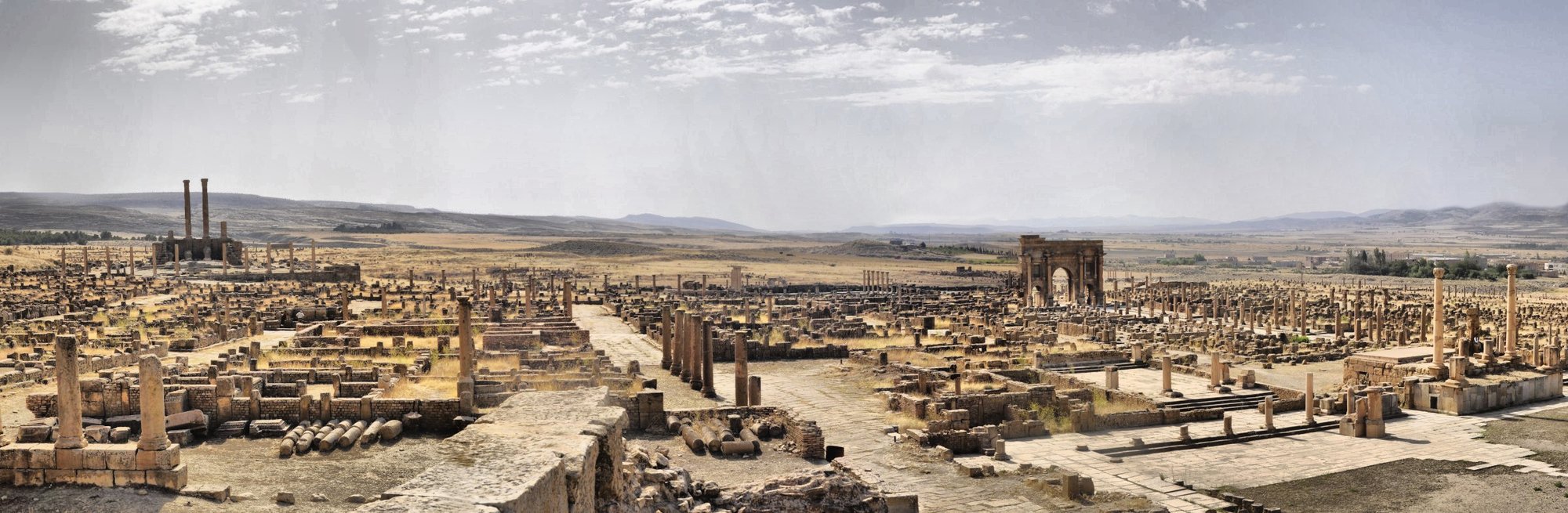 A wide colour panorama of Timgad looking across the excavated city, with the columns of the Capitol rising at the far left, the colonnaded decumanus maximus running through the centre, the Arch of Trajan standing to the right, and the Aurès Mountains on the horizon.
