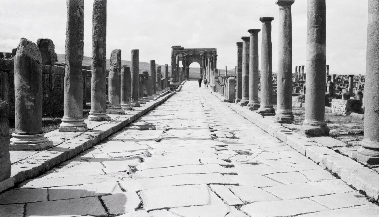 A black and white photograph of Timgad's colonnaded main street, worn paving stones stretching toward the Arch of Trajan in the distance
